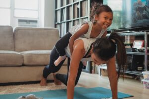 Young Asian woman piggybacking smiling daughter while exercising at home